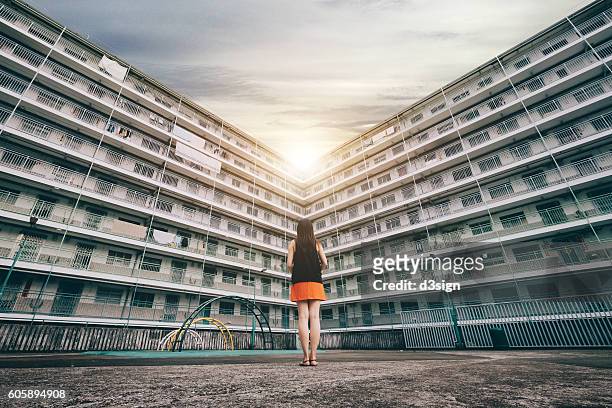 the rear view of woman standing against old apartment building, looking towards the sun shining - social housing stock pictures, royalty-free photos & images