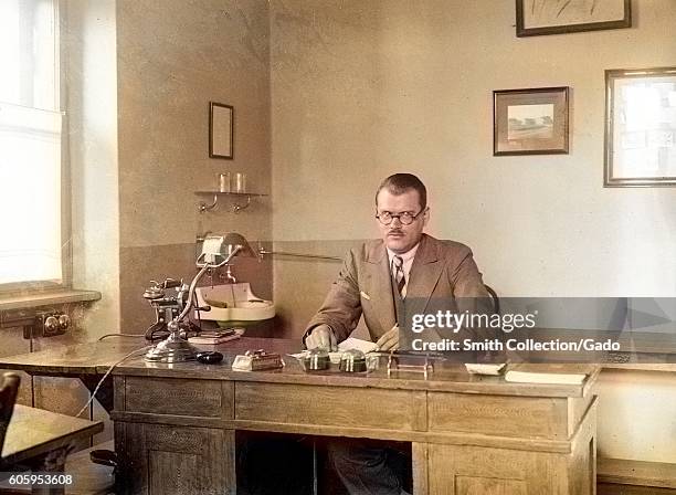 Man with short hair, round spectacles, a conservative suit and a stern facial expression sits behind a large wooden desk in his office, with a small...