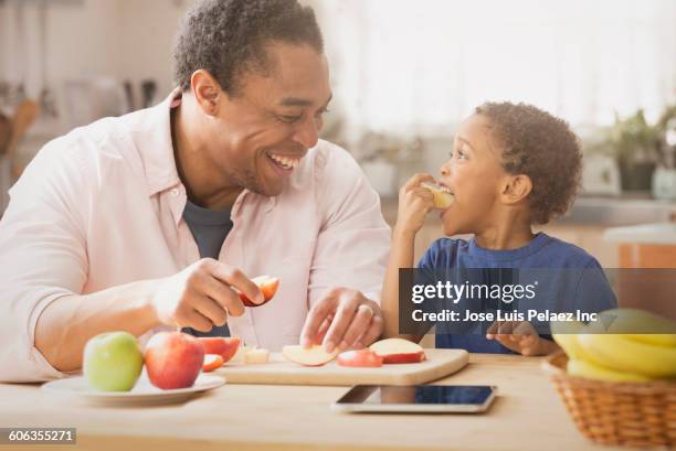 mixed race father and son eating apples in kitchen - child holding apples stock-fotos und bilder