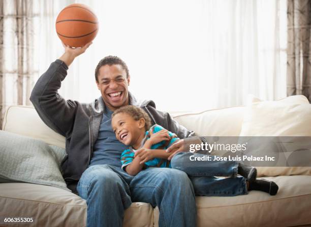 mixed race father and son playing with basketball on sofa - african family watching tv stockfoto's en -beelden