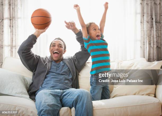 mixed race father and son cheering with basketball on sofa - basketball fans stock pictures, royalty-free photos & images
