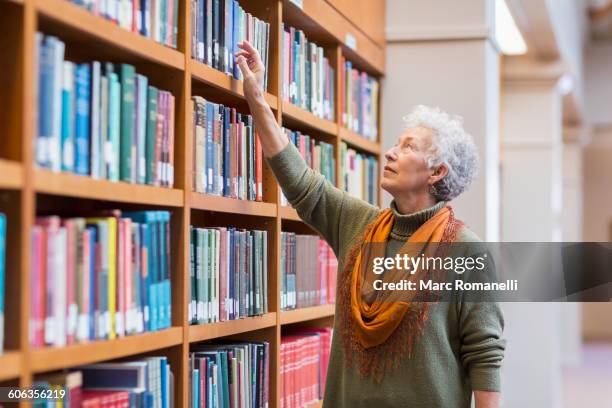 older mixed race woman choosing book in library - differential focus education reach stockfoto's en -beelden