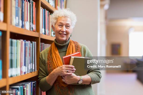older mixed race woman holding books in library - senior women india fotografías e imágenes de stock