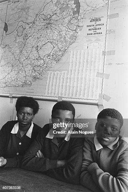 Three students sit under a political map of South Africa showing the "homelands" or zones reserved for blacks. | Location: Crossroad, South Africa.