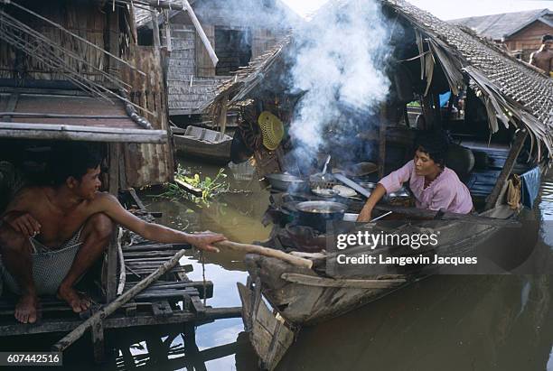 Woman prepares food on a boat belayed to a floating bungalow where a man sits, in the lake village of Phtout, on Tonle Sap Lake. | Location: Phtout,...