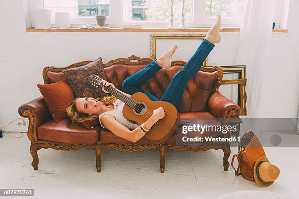 happy woman lying on leather couch playing guitar - blonde long legs fotografías e imágenes de stock