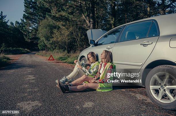 two young women with safety vests sitting in the road awaiting assistance for their damaged vehicle - vehicle breakdown ストックフォトと画像