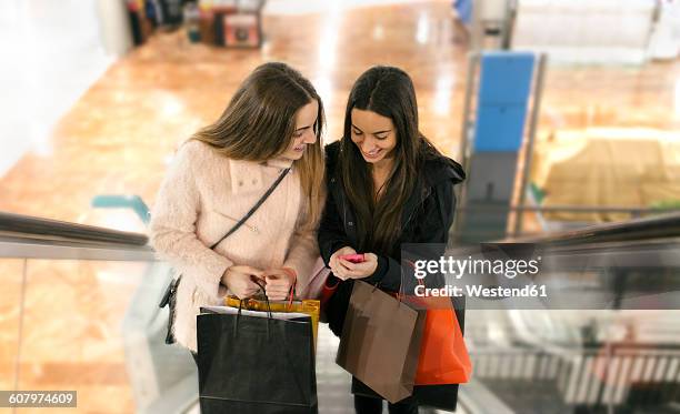 two female friends with shopping bags on an escalator of a shopping center looking at smartphone - girl after shopping stock-fotos und bilder