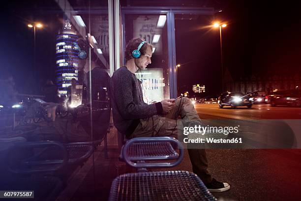 germany, munich, man with headphones sitting at bus stop using digital tablet at night - bus stop stock pictures, royalty-free photos & images