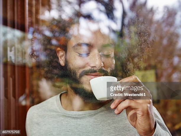 smiling young man enjoying cup of coffee behind windowpane - indulgence foto e immagini stock