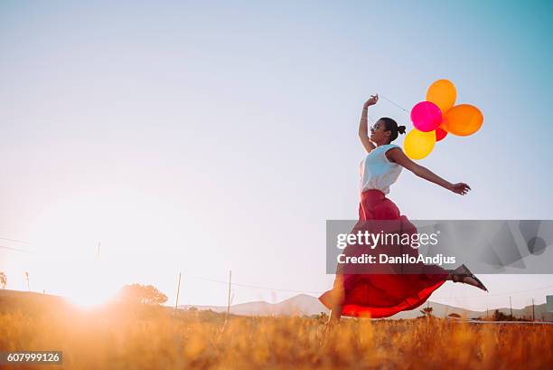 young woman running threw the fields holding balloons - multi colored skirt stockfoto's en -beelden