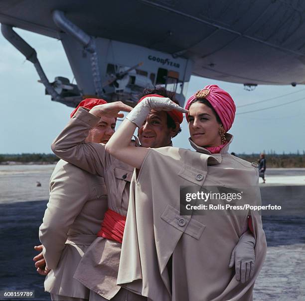 From left to right, actors Leo McKern , John Bluthal and Eleanor Bron posed together in costume in front of a Goodyear blimp during filming of the...