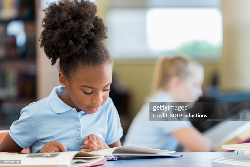 Cute African American schoolgirl reading a book in the library