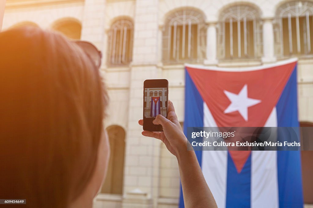 Over the shoulder view of young woman photographing Cuban flag on smartphone, Havana, Cuba