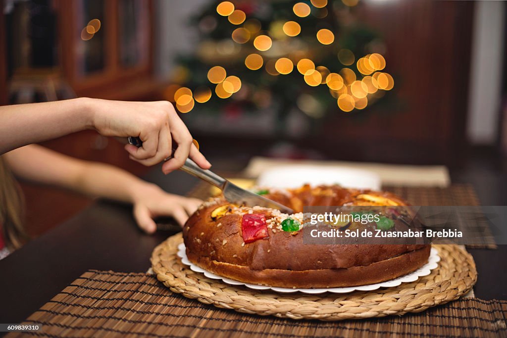 Hands of a girl cutting a Christmas fruitcake besides the Christmas tree.