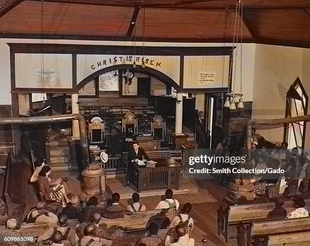 African-American Farm Security Administration borrowers sitting in a church and listening to an official, with a message above the altar reading...