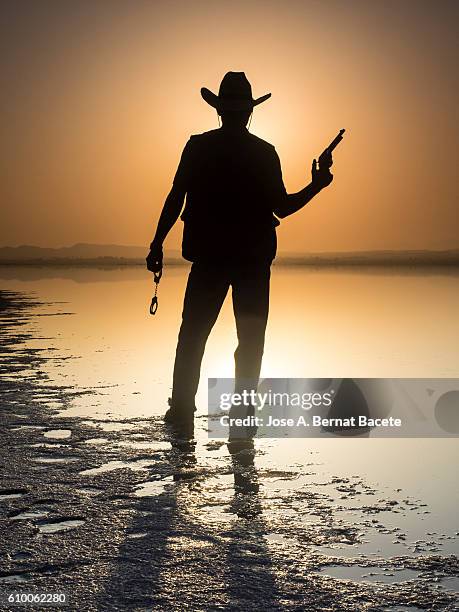 silhouette of a man with cowboy clothes and pistoala front of a lake - modern cowboy stock-fotos und bilder