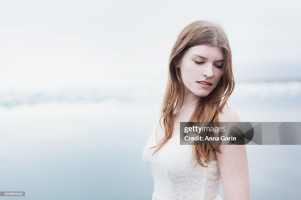 Headshot of beautiful young woman in sleeveless white lace dress, looking down, bright outdoor background
