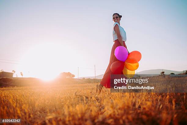 cheerful beautiful young woman in the field holding colorful balloons - multi colored skirt stockfoto's en -beelden