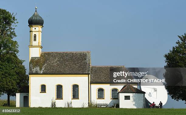 Bikers make a break in front of a parabolic antenna and the Baroque church of Saint John in Raisting near Weilheim, southern Germany, during nice...