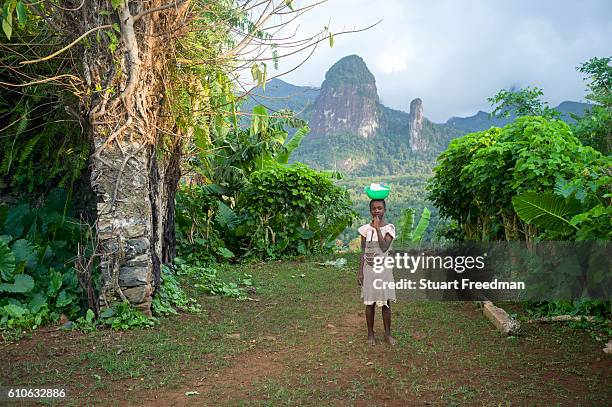 Child carrying a bowl on her head stands in from of the Pico de Príncipe and the Pico Cão Grande, the two highest mountains on the island, Principe,...