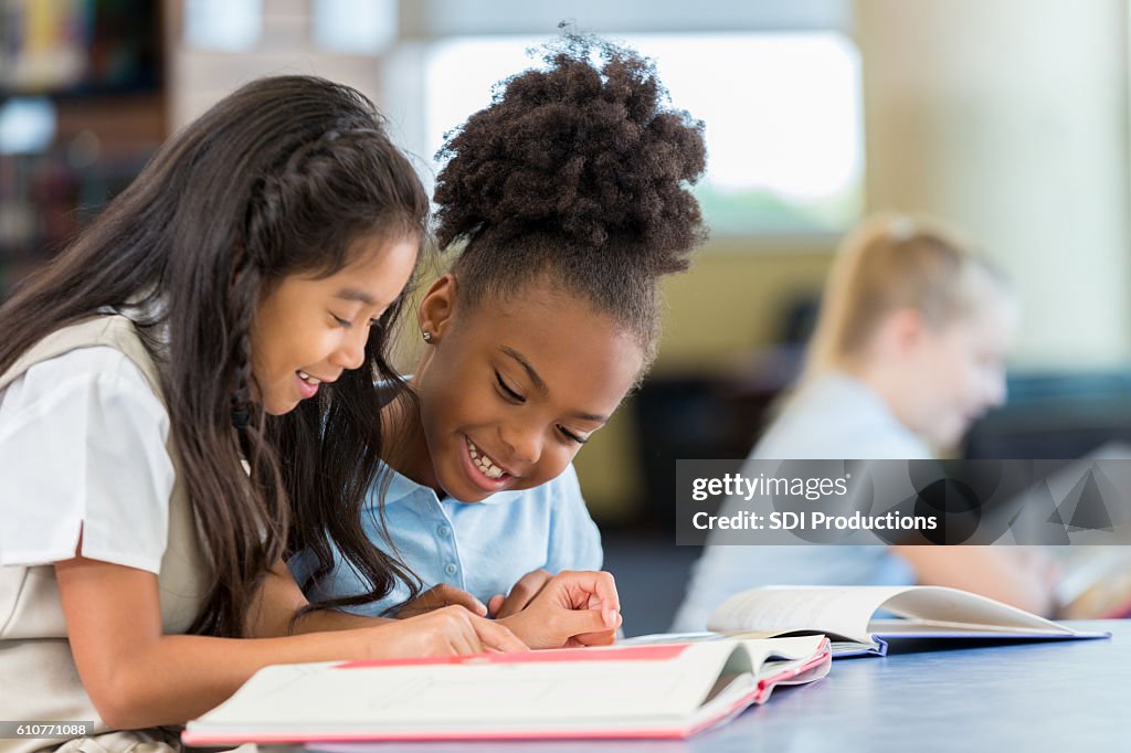 Smiling and cheerful schoolgirls reading a book together at school