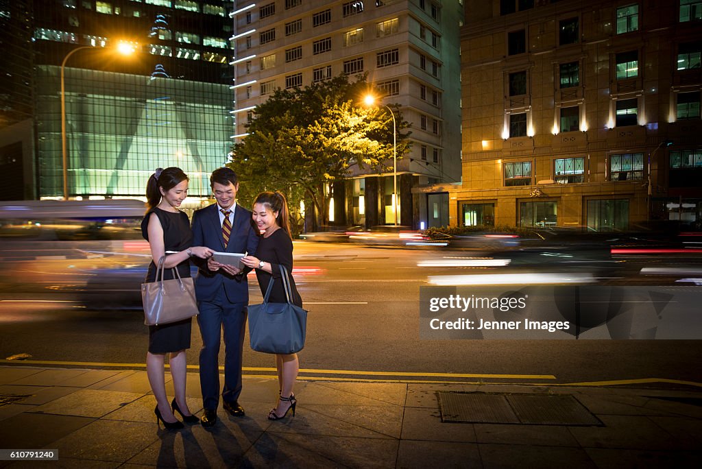 Three Asian business collegues looking at a Tablet device at night with traffic light trails in background.