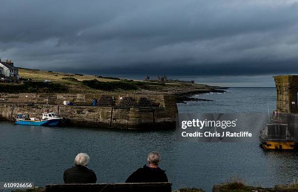 Two men sit on a bench by the fishing port of Craster as dark storm clouds form over Dunstanburgh Castle in the distance on the coast of...