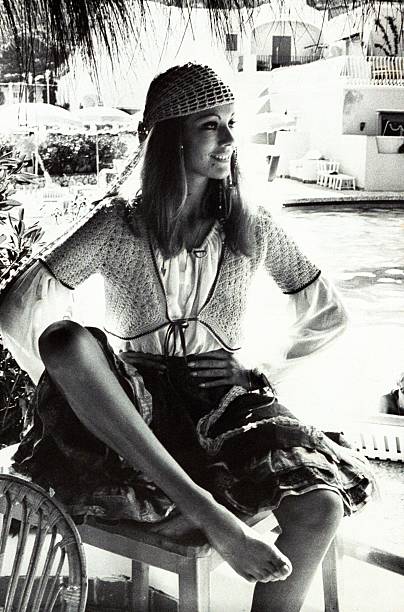 Claudine Auger wearing a short crocheted vest over a drawstring blouse, crocheted skull cap and a melange of Indian cotton skirts; seated beside a...