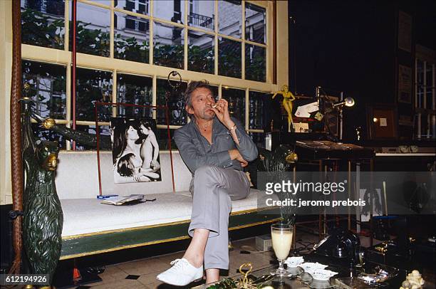 French singer and songwriter Serge Gainsbourg in his Paris home located on Rue de Verneuil. Next to him is a picture of his wife Jane Birkin and...