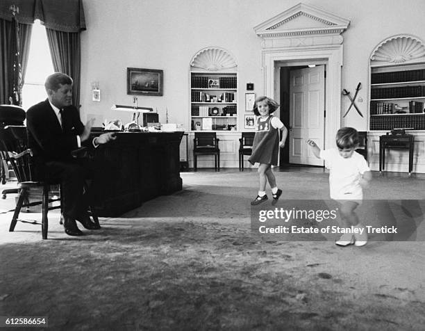 American President John F. Kennedy claping while watching his daughter, Caroline, and son, John Jr., dance in the Oval Office.