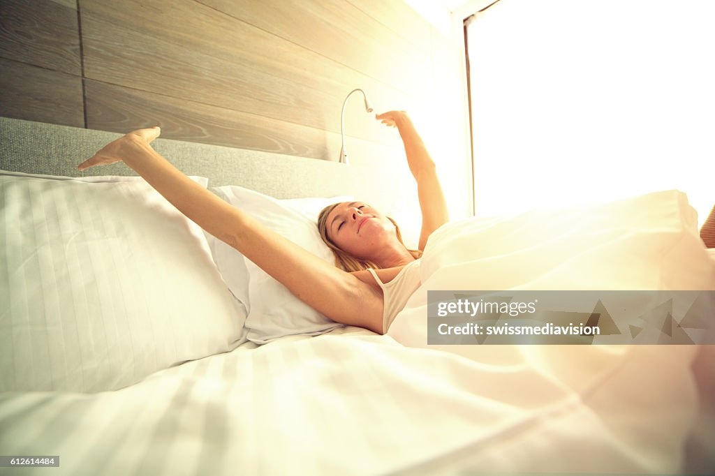 Young woman waking up in her hotel room, stretching arms