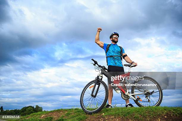 glücklicher junger biker steht hand oben auf dem berg - bike headset stock-fotos und bilder