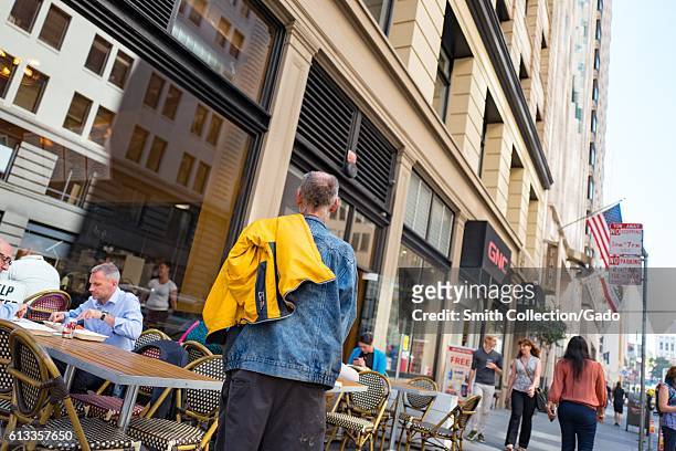 View of a homeless man, in a denim jacket, near a sidewalk restaurant where a man in a button down shirt eats lunch, in the Financial District...