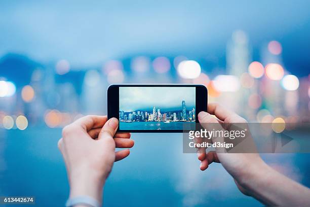 human hands taking pictures with smartphone over hong kong cityscape and victoria harbour at sunset - fotoberichten stockfoto's en -beelden