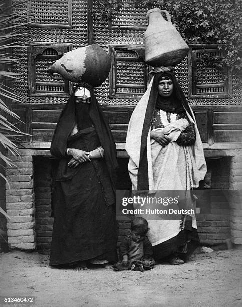 Baby plays on the ground at the feet of two Arab women who are balancing large pottery jars on their heads in Egypt.