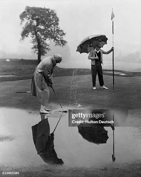 Playing barefoot, George Doonan puts the golf ball on a rain-soaked green while Roy Bentley holds the flag during practice before a golf match...