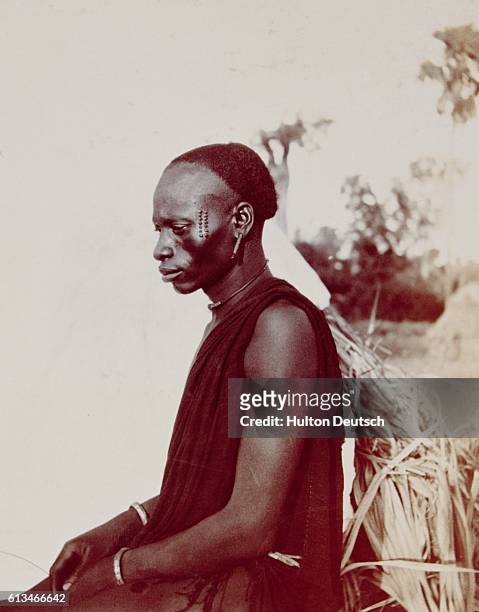 African Man in Rural Setting. Half body profile shot of African man wearing material draped over shoulder, earrings, choker and bracelets. He has...