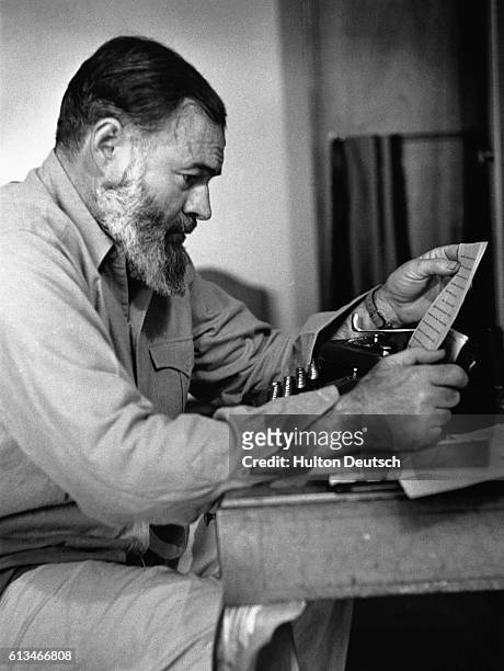 Ernest Hemingway peruses a page of his writing from a typewriter at his desk.