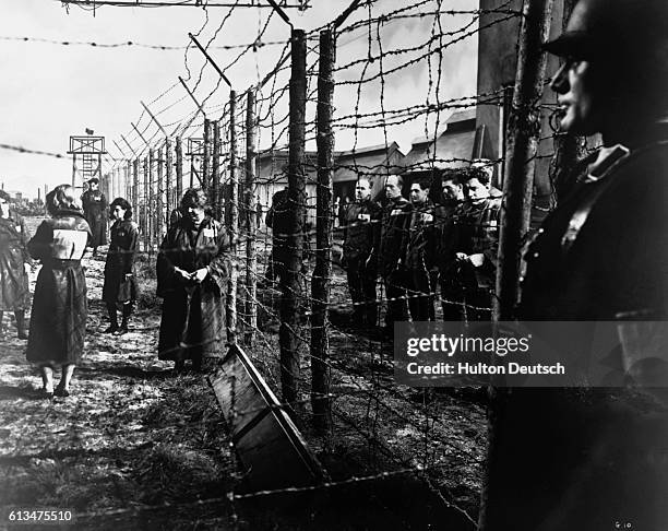 An electrified barbed-wire fence separates male and female prisoners at a German concentration camp. A Nazi guard keeps watch in the foreground. The...