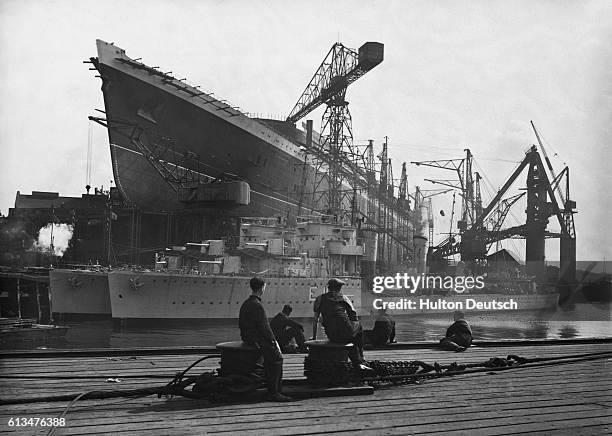 The new Cunard White Star liner RMS Queen Elizabeth, under construction, dwarfs two Argentine destroyers at the shipyard of John Brown & Co. Ltd. In...