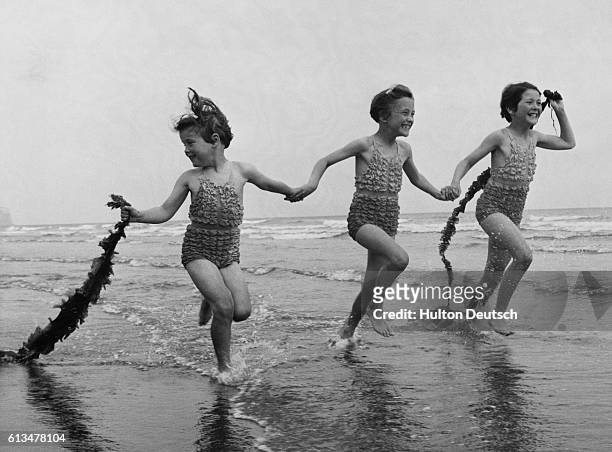Elaine, Veronica, and Christine Patrickson , aged 6 and 9, run through the surf, trailing tails of seaweed behind them.