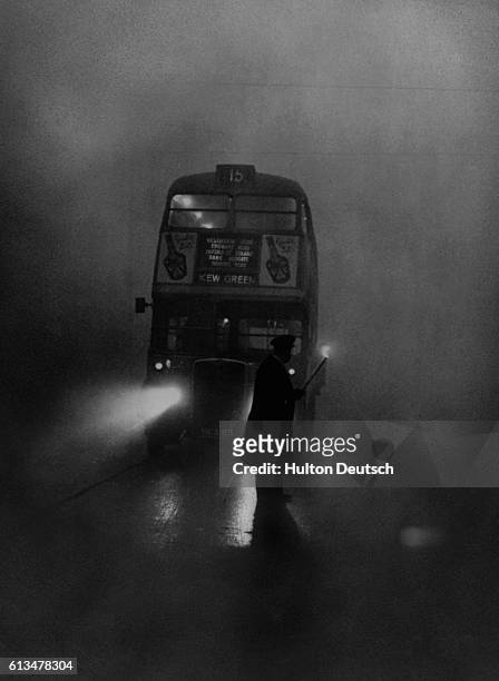 Bus inspector uses a flare to lead a London double-decker bus over a crossing in the fog.