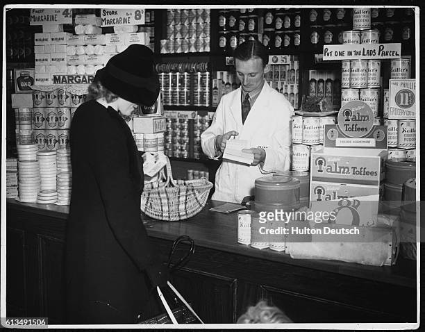 London housewife places her butter rations into her basket while the grocer snips out the corresponding coupon from the ration book.
