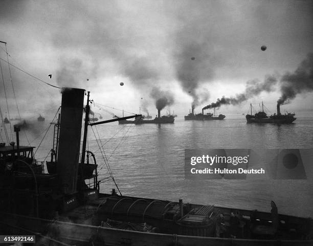 Coastal convoy prepare to leave a British port at dawn guarded by escord ships, an RAF umbrella and barrage balloons which can be seen through the...