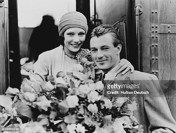 Movie stars Gary Cooper and Lupe Velez, dating at the time, hold a large bouquet of roses together at a train car in Hollywood.