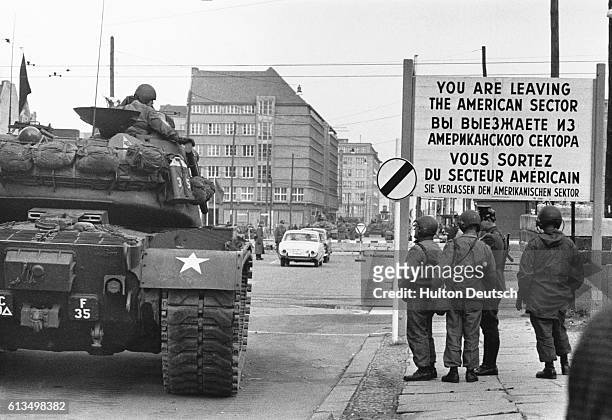 American soldiers and a tank near one of the checkpoints of the Berlin Wall. A sign in English, French, Russian and German warns that "You are...