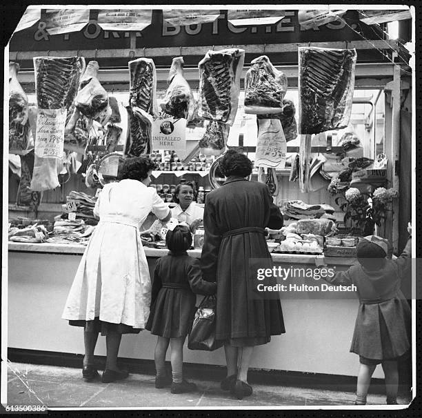 Mother makes a purchase at a meat counter while one of her children investigates something nearby which has caught his eye.