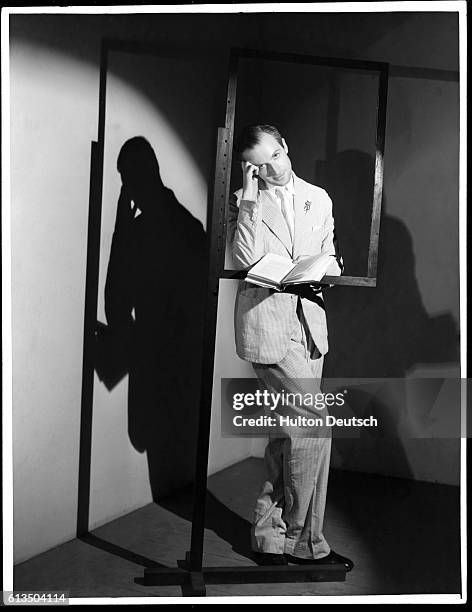 English photographer and set designer Cecil Beaton leans against a frame on a stand while reading a book.