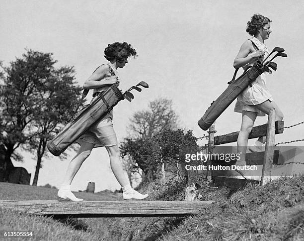 Two young women, both sporting short-sleeved tops and shorts, carry their golf bags across a bridged ditch and stile on the Filton course, at...